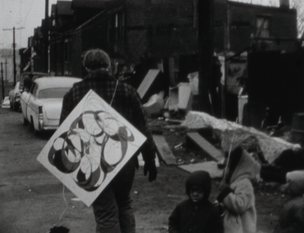A black and white image of the back of a man walking through a city street littered with trash. He has a painting slung over his shoulder. To the right of him are two Black children, one holding an umbrella and the other looking directly into the camera lens.
