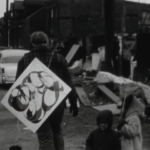 A black and white image of the back of a man walking through a city street littered with trash. He has a painting slung over his shoulder. To the right of him are two Black children, one holding an umbrella and the other looking directly into the camera lens.
