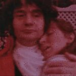 A color film still of a man of a cravat-wearing man embracing a woman.