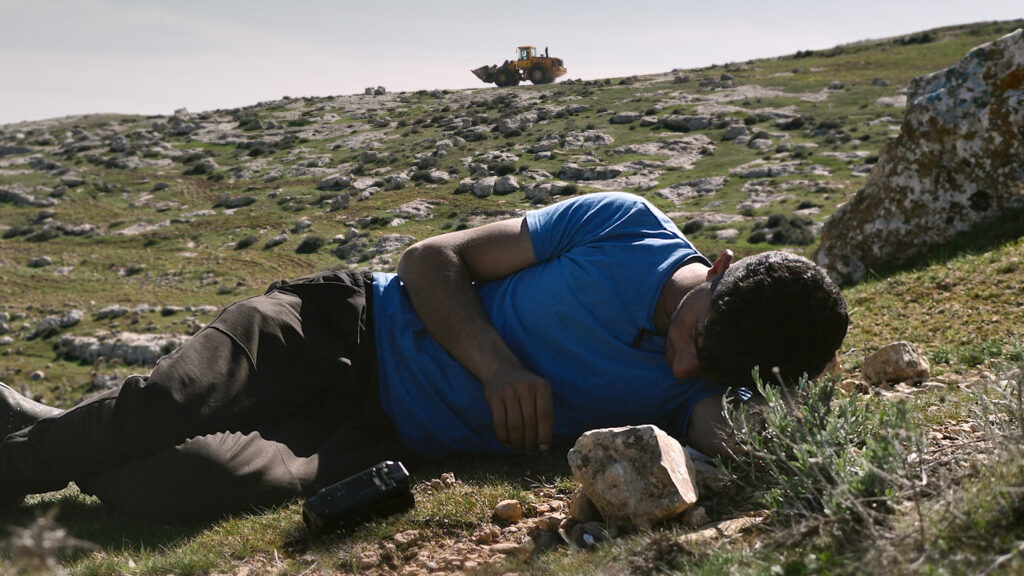A color film still of a man lying on his side on a rocky green hillside, looking forlornly down at the grass. A camcorder sits on the ground just before him. Far in the distance behind him, a bulldozer drives down the hill.
