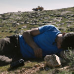 A color film still of a man lying on his side on a rocky green hillside, looking forlornly down at the grass. A camcorder sits on the ground just before him. Far in the distance behind him, a bulldozer drives down the hill.