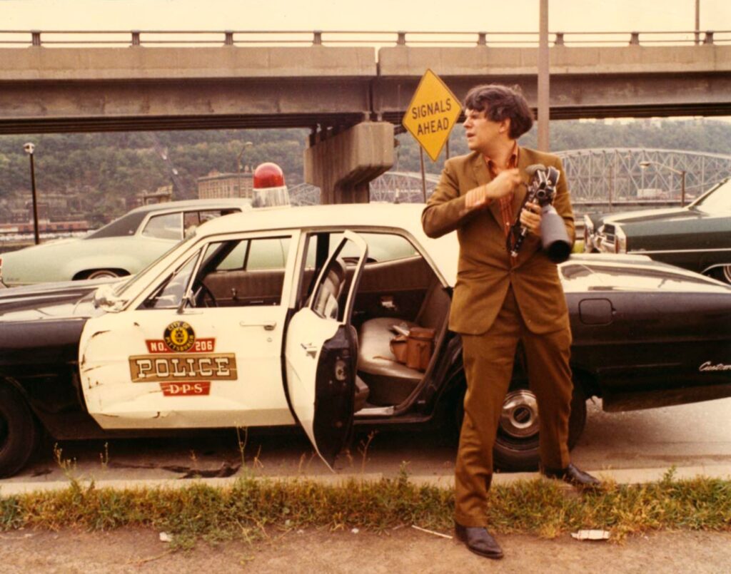 A photo of a man (Stan Brakhage) wearing a brown suit, holding a 16mm camera with a zoom lens to his chest, and looking to his right as if examining something or urgent interest. He is standing in front of a police car with the back door open. A highway overpass and city hills are visible in the background.