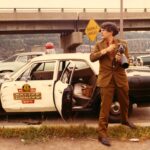 A photo of a man (Stan Brakhage) wearing a brown suit, holding a 16mm camera with a zoom lens to his chest, and looking to his right as if examining something or urgent interest. He is standing in front of a police car with the back door open. A highway overpass and city hills are visible in the background.