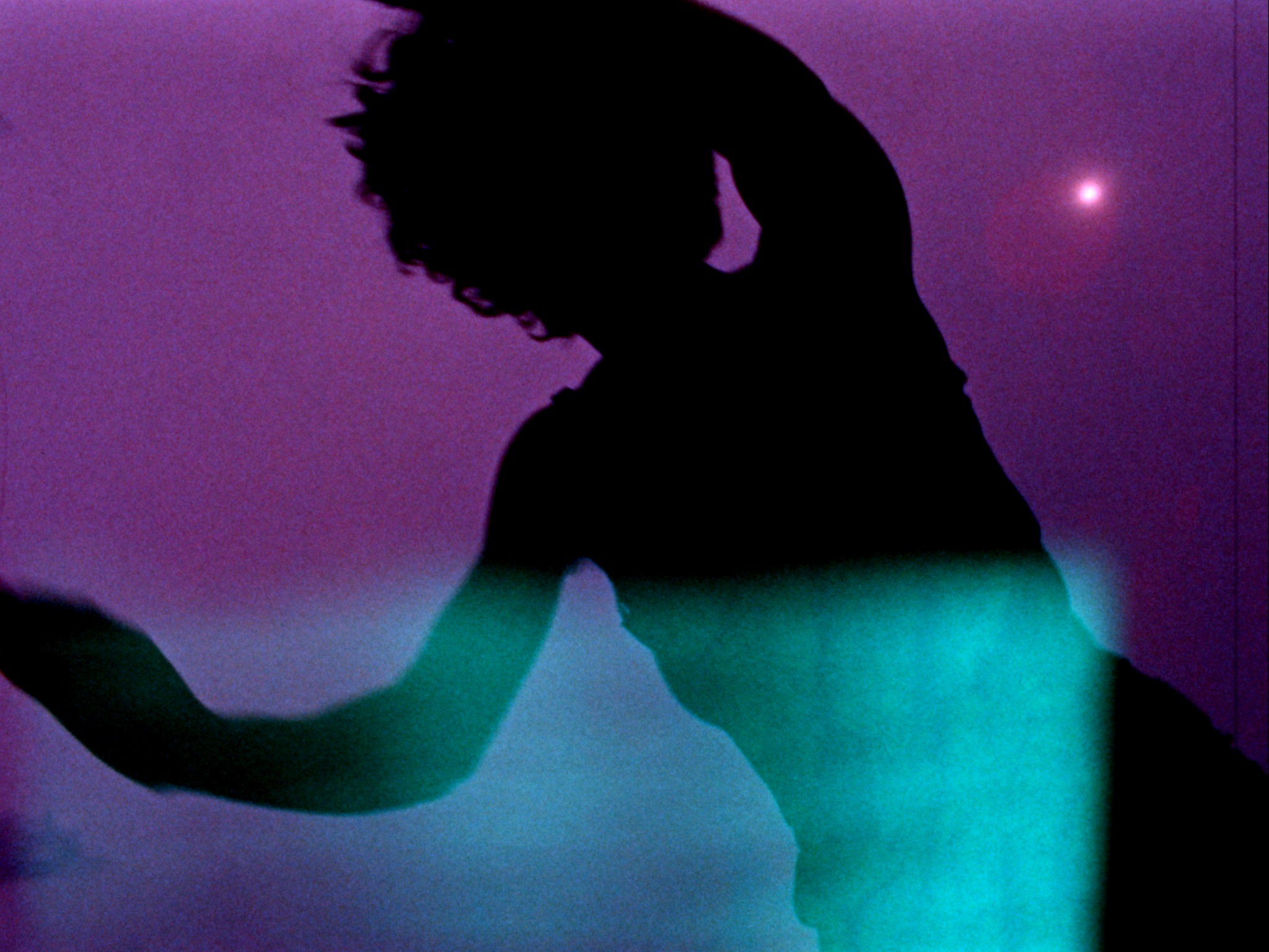 A color film still of the silhouette of a person with arms raised above their head, as if they're floating across a purple-ish landscape.