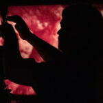 A color photograph of the silhouette of a projectionist threading up a 16mm projector in front of a screen with a red image on it.
