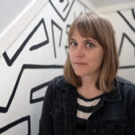 A color photograph of Lori Felker sitting in a white staircase with black, freeform lines adorning the walls.