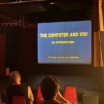 A color photograph of the backs of heads of people in a movie theater audience. Projected on screen is a title card for a movie called "The Computer and You."