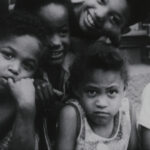 A black and white film still of four Black children pressed together on a city street, posing for the camera.