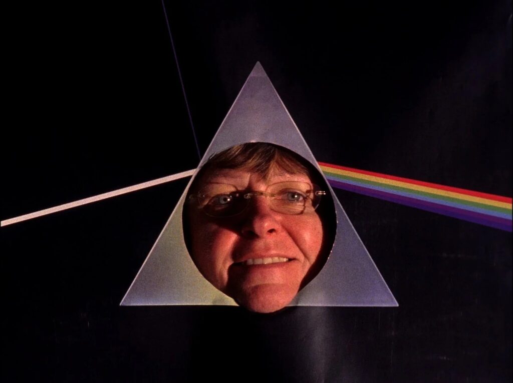 A color photograph of a white woman in her 50s or 60s smiling with her face sticking through a circular cutout in the middle of the triangle from the cover of Pink Floyd's Dark Side of the Moon