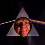 A color photograph of a white woman in her 50s or 60s smiling with her face sticking through a circular cutout in the middle of the triangle from the cover of Pink Floyd's Dark Side of the Moon