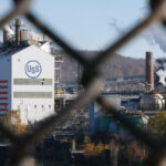 A color movie still (16:9 aspect ratio) with a chain link fence slightly our of focus in the extreme foreground. In the distance is a modern steel mill with the US flag painted on one side and the USS Steel logo on another. To the right are smokestacks discharging billowing clouds of white smoke. Gentle Appalachian hills are further in the distance